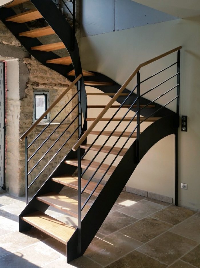 Escalier deux quarts tournant à limon en tôle noire, marches de chêne, garde-corps à lisses et main courante bois, contre un mur en pierre